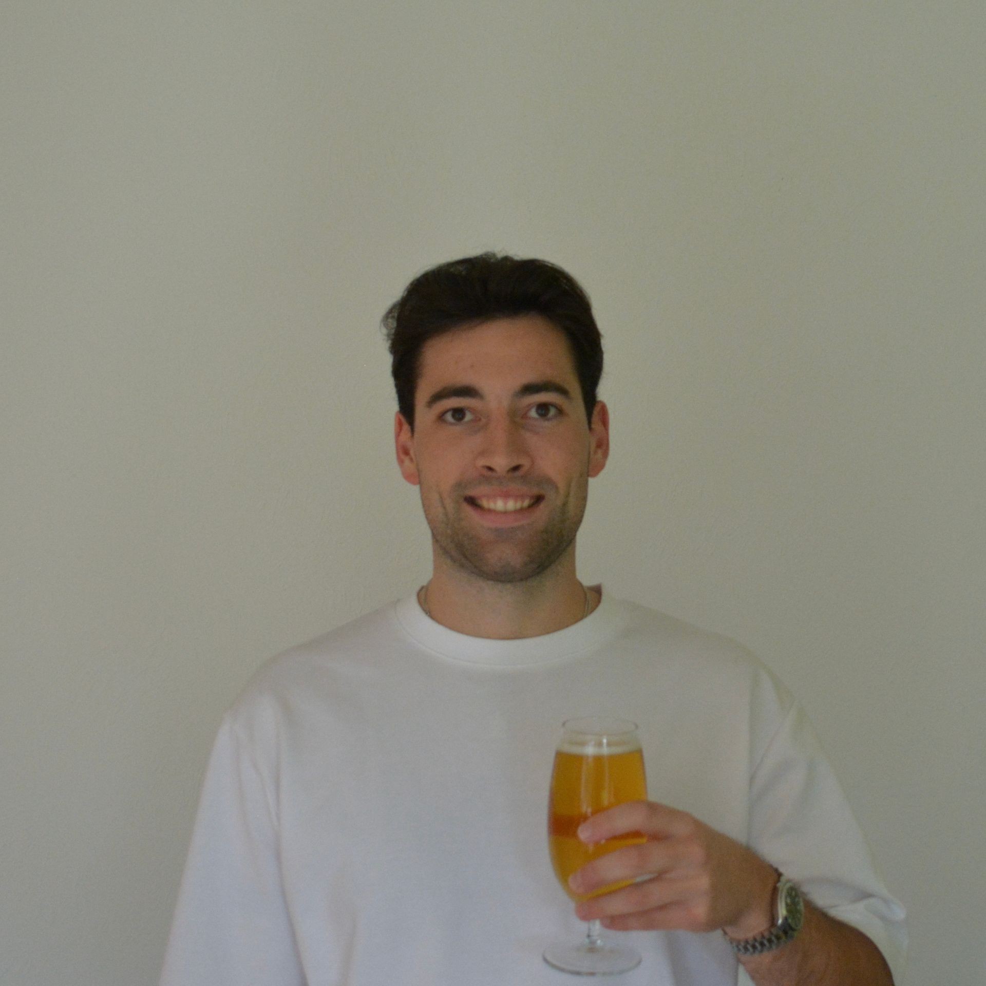 Person wearing a white shirt holding a glass of orange beverage next to a plain wall.