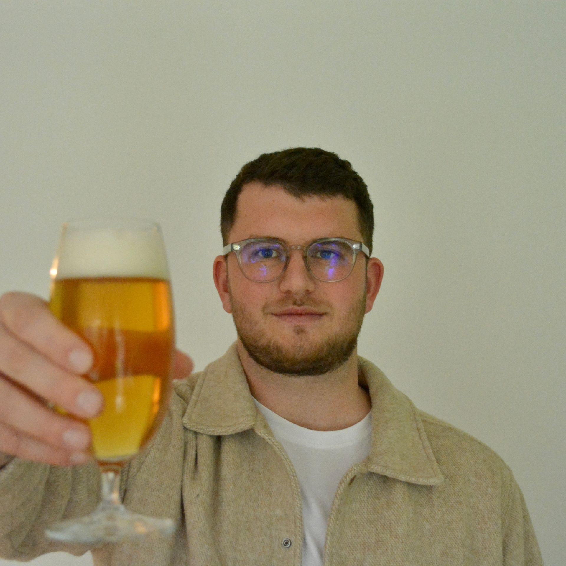 Person holding up a glass of beer with frothy head against a plain background.